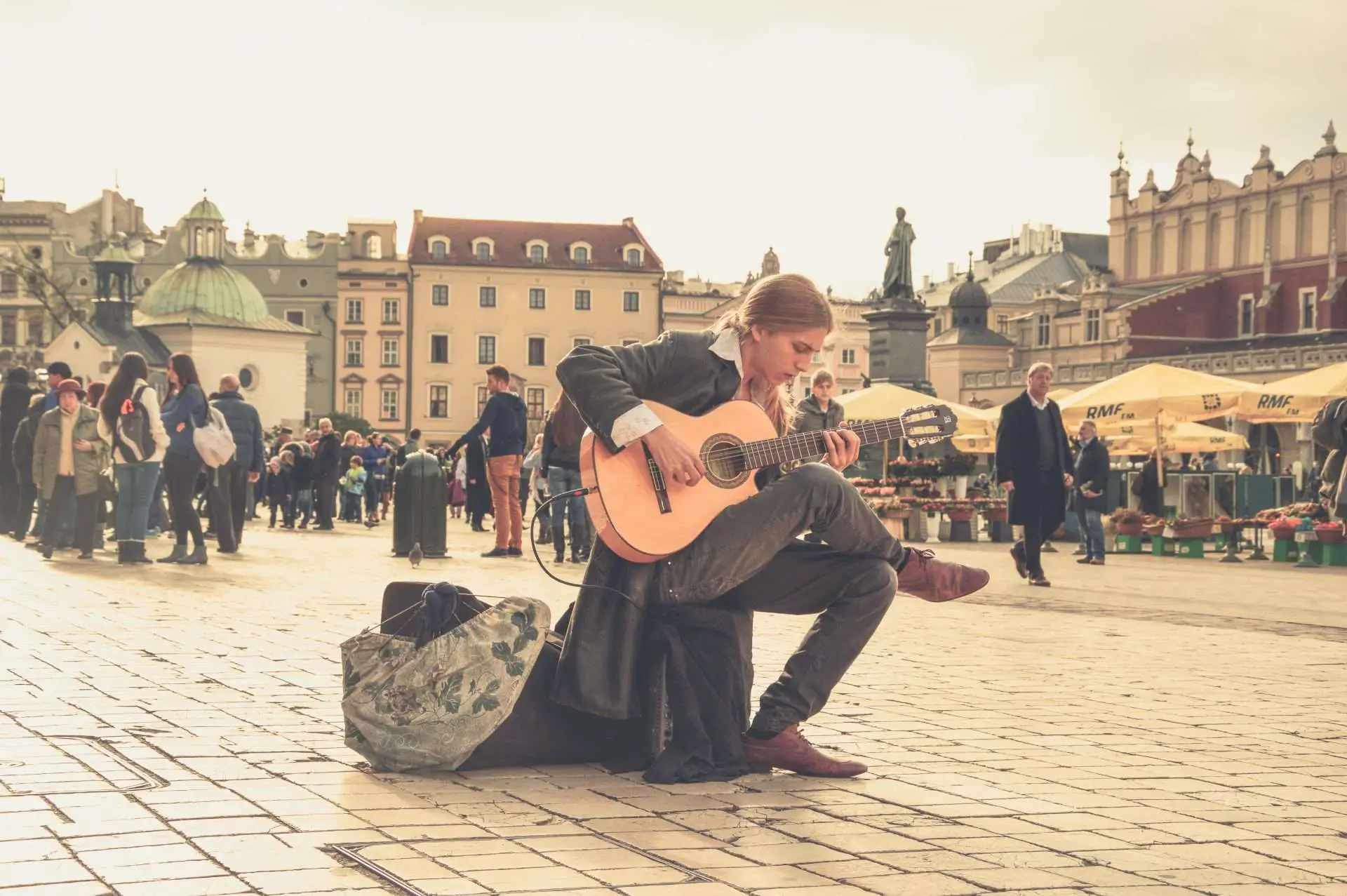 man sitting down at a town square and playing his guitar with people walking by