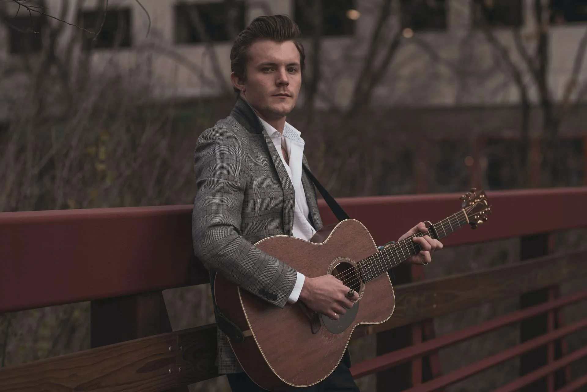 man leaning against a fence & holding an acoustic guitar